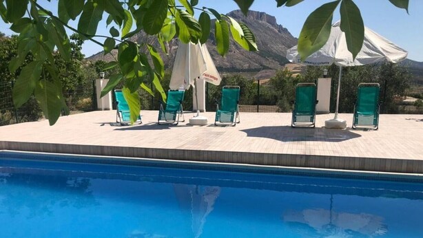 A swimming pool with lounge chairs and umbrellas, surrounded by a wooden deck and a mountainous backdrop.