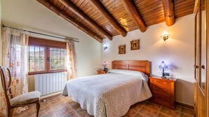 A bedroom with a wooden ceiling, a bed with a headboard, a nightstand, a chair, a window with curtains, and wall-mounted lights.