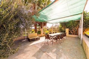 Une terrasse extérieure avec une table et des chaises, entourée de plantes et dotée d’un espace ombragé.