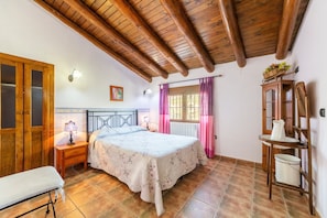 Une chambre avec un plafond en bois, un lit recouvert d’une couvre-lit à motifs floraux, une table de chevet avec une lampe, une fenêtre avec des rideaux et une armoire en bois.