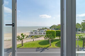 Vista a través de una ventana de una zona costera con playa, un edificio de diseño moderno y abundante vegetación.