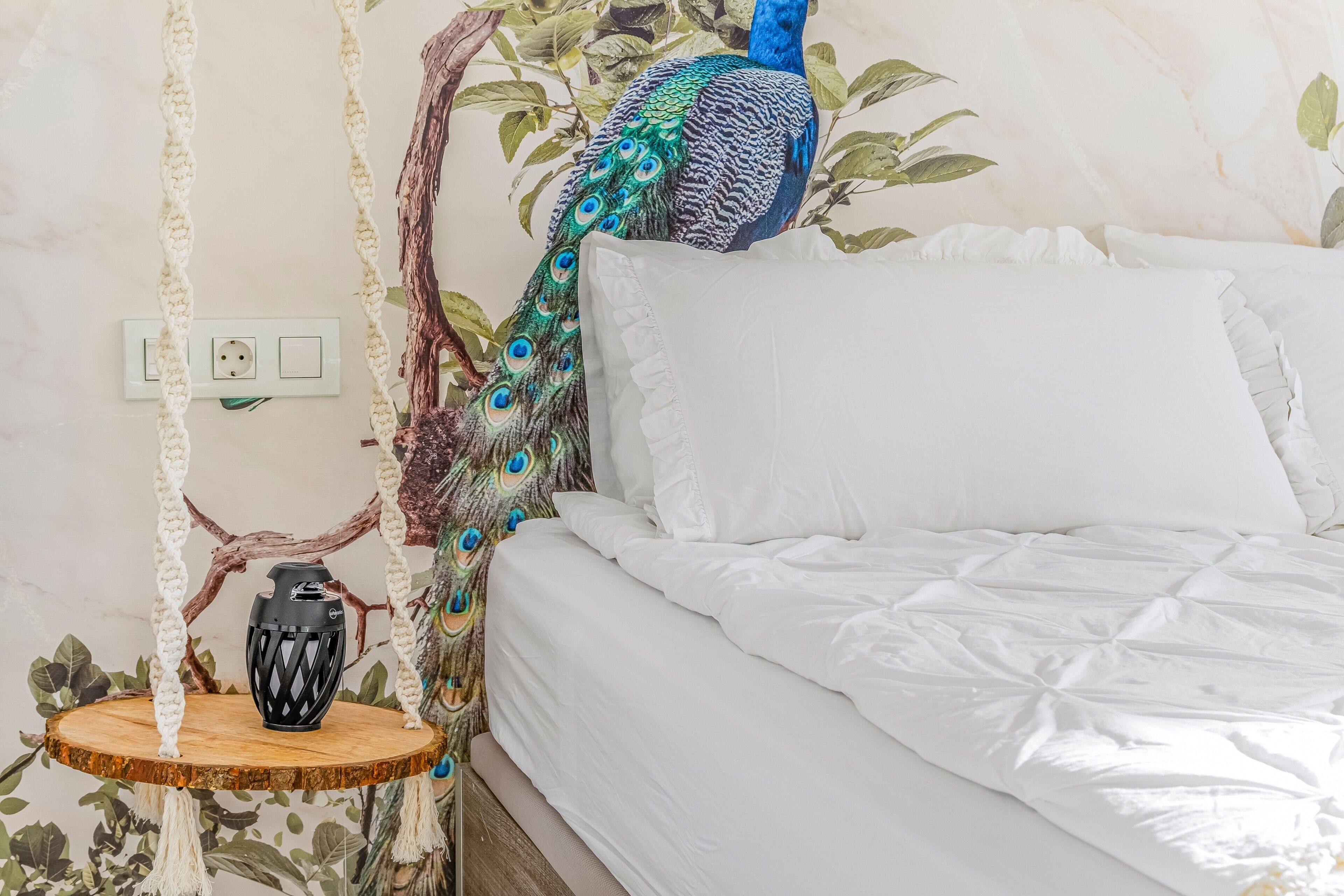 Un lit avec une literie blanche, un fauteuil suspendu en macramé et un paon décoratif.