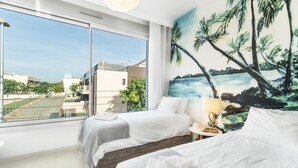 Een hotelkamer met twee bedden, een groot raam, een schilderij van palmbomen en uitzicht op gebouwen.