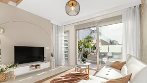Een moderne woonkamer met een flatscreen televisie, een witte bank en een groot raam met uitzicht op gebouwen.