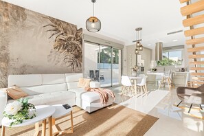 A modern living room with a white sofa, a coffee table, a dining area with a white table and chairs, a kitchen in the background, and a large window providing a view.