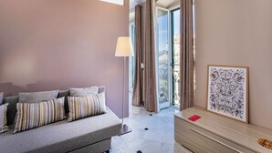 A modern hotel room with a grey sofa, striped pillows, a wooden side table with a framed picture, and a view of the city through large windows with curtains. - City and the Sea by Wonderful Italy (Genoa)