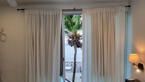 Quarto de hotel com cama, cortinas, ventilador de teto, janela com vista para palmeiras e praia, e mesa de cabeceira com abajur.