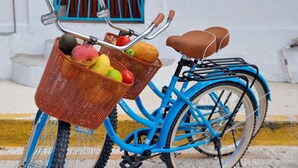 Uma bicicleta azul com cesta contendo frutas e legumes, estacionada em frente a um edifício branco com porta azul e placa com o número 2.