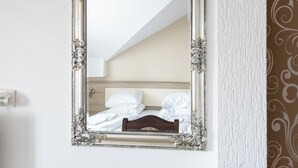 Ein Zimmer mit einem Bett, einem Holzstuhl und einem Spiegel mit aufwendiger Rahmung an der Wand.