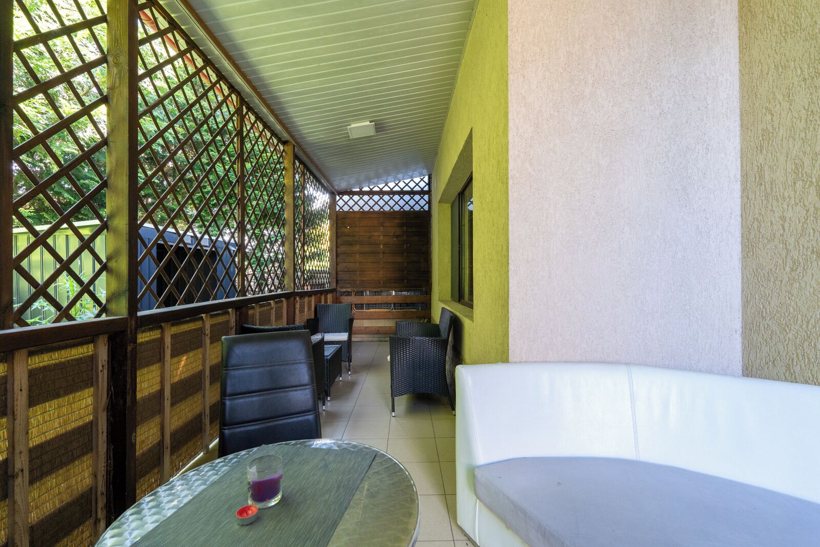 Un espacio de estar al aire libre cubierto, con mobiliario de mimbre y un sofá blanco, delimitado por celosías de madera y una pared amarilla.