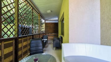 Un espacio de estar al aire libre cubierto, con mobiliario de mimbre y un sofá blanco, delimitado por celosías de madera y una pared amarilla.