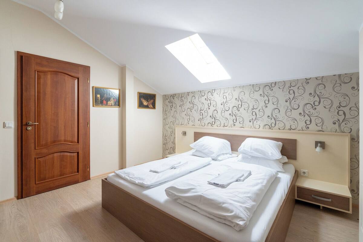Un dormitorio con puerta de madera, cama con ropa de cama blanca, tragaluz y papel tapiz estampado.