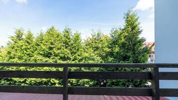 A balcony with a wooden railing, a red roof, and dense green trees in the background.