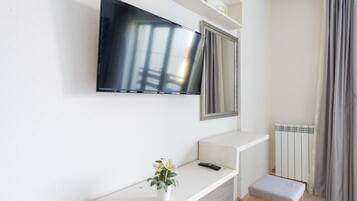 Una habitación moderna con un televisor de pantalla plana montado sobre un mueble blanco, un espejo, una planta en maceta, un control remoto y un banco.
