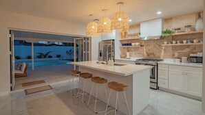 Una cocina moderna con una isla grande, taburetes de bar, luces colgantes y vista a una piscina.