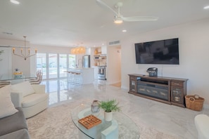 A modern living room with a glass coffee table, a sofa, a TV mounted on the wall, and a kitchen in the background.