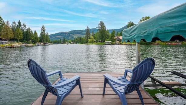 A wooden dock with two blue chairs facing a calm lake, surrounded by trees and a distant mountain range.