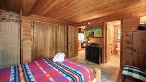 Une pièce de cabane en bois comprenant un lit, une télévision, un bureau et une salle de bain, visible à travers une porte ouverte.