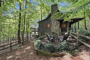 A cabin with a stone chimney, wooden deck, and outdoor seating area surrounded by trees. - The Guest House Historic Cabin in Big Canoe (Jasper)