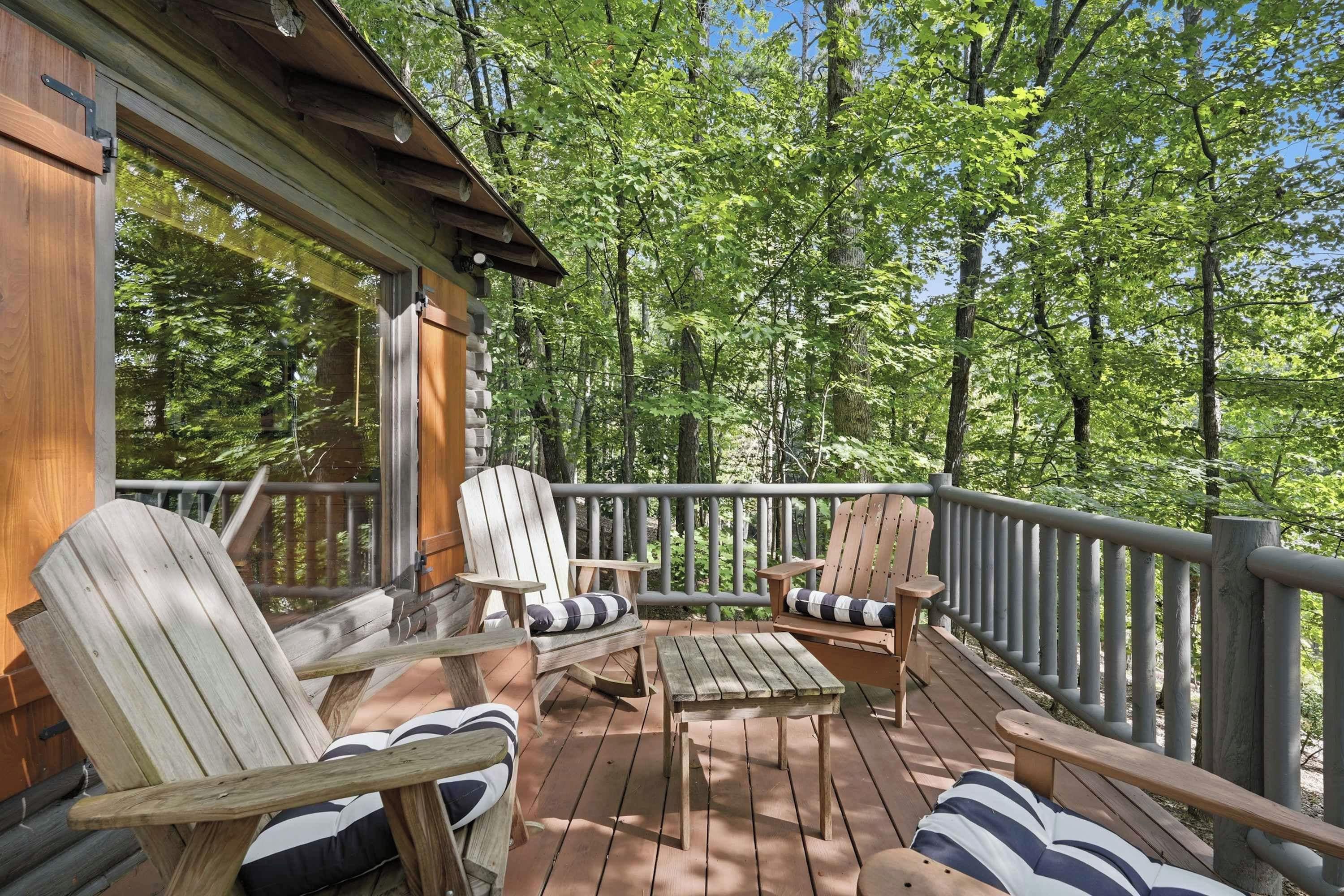A wooden deck with Adirondack chairs and a table, surrounded by a forest.