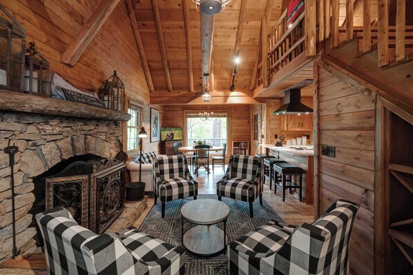 A cabin interior with a stone fireplace, checkered sofas, a round table, and a dining area with chairs. - The Guest House Historic Cabin in Big Canoe (Jasper)
