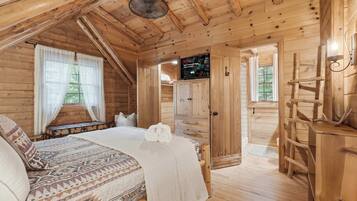 A cozy wooden cabin bedroom with a bed, a window with curtains, a TV mounted on the wall, and a ladder against the wall.