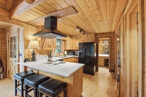 A wooden cabin interior with a kitchen featuring a central island, bar stools, a stove, and a refrigerator. - The Guest House Historic Cabin in Big Canoe (Jasper)
