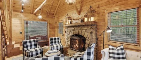 Interior acogedor de una cabaña de troncos con chimenea de piedra, muebles a cuadros y ventanas amplias.