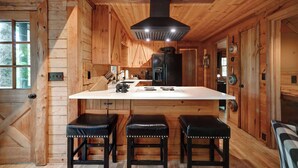 L’intérieur d’un chalet en bois comprend un espace cuisine avec un plan de travail blanc, des tabourets noirs et une hotte noire.