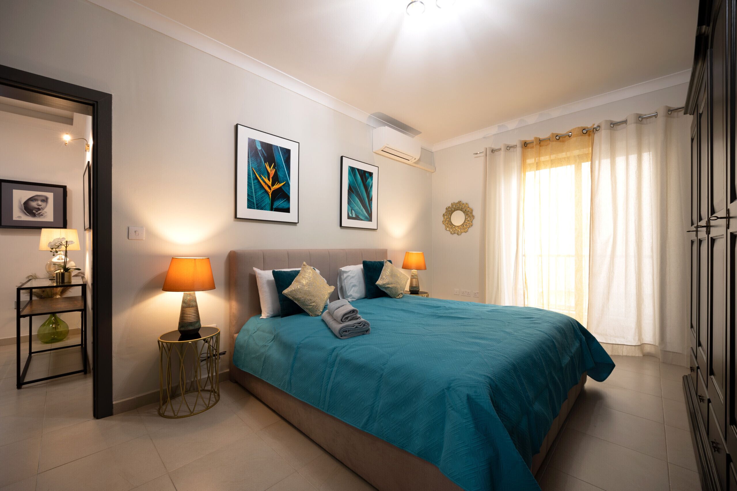 A bedroom with a bed, two bedside lamps, a wall-mounted air conditioner, and a window with curtains.