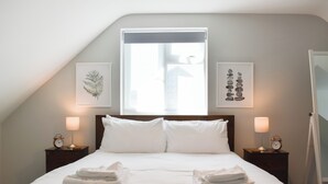 Een keurig opgemaakt bed met witte lakens en handdoeken, twee nachtkastjes met lampen, en ingelijste kunstwerken aan de wanden.
