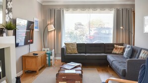 Een moderne woonkamer met een grijze bank, een houten salontafel, een vloerlamp en een aan de muur bevestigde televisie. Er staan koffers op de vloer, wat duidt op een recente reis.