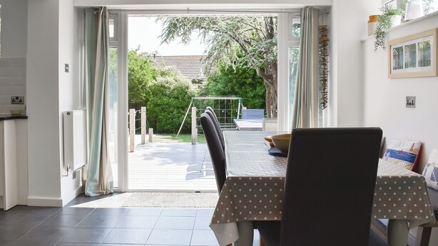 Een eetruimte met een tafel en stoelen, uitzicht op de tuin door een open deur, en een wit plafond met inbouwverlichting.