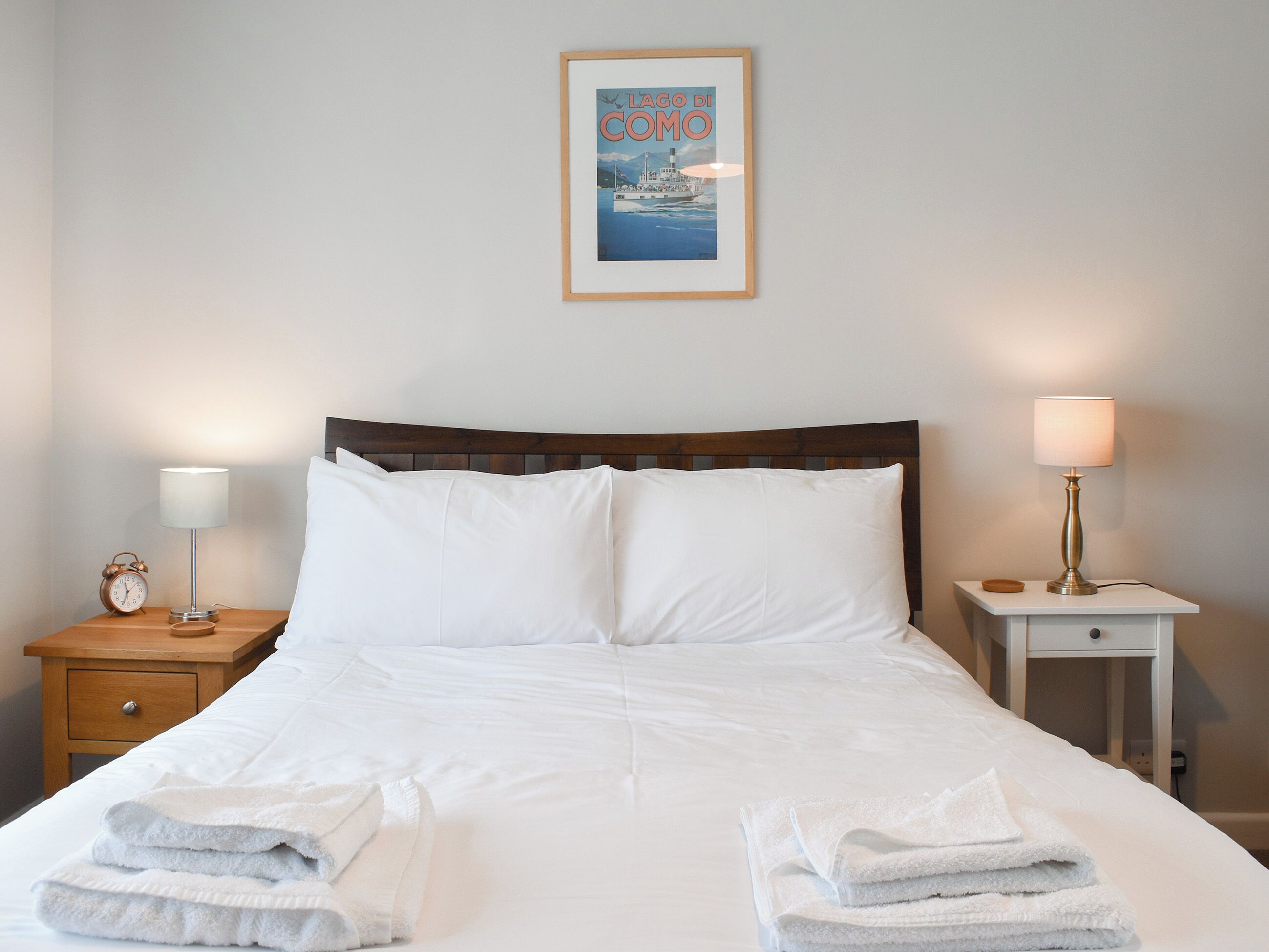 Een net opgemaakt bed met witte lakens en handdoeken, een ingelijst beeld erboven en nachtkastjes met lampen.