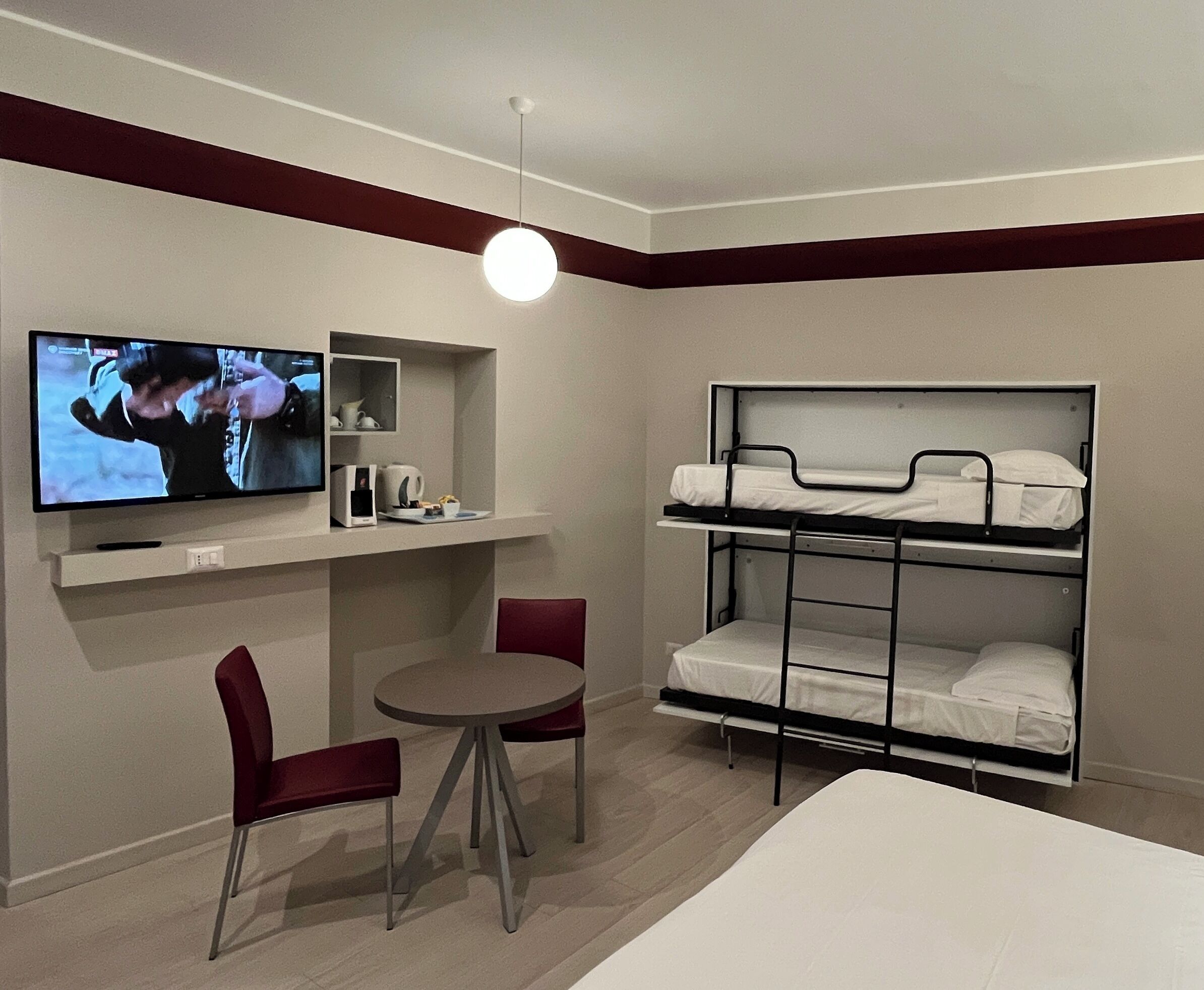 Ett hotellrum med en våningssäng, en tv, ett litet bord med två stolar och en hylla med en kaffebryggare och andra föremål.