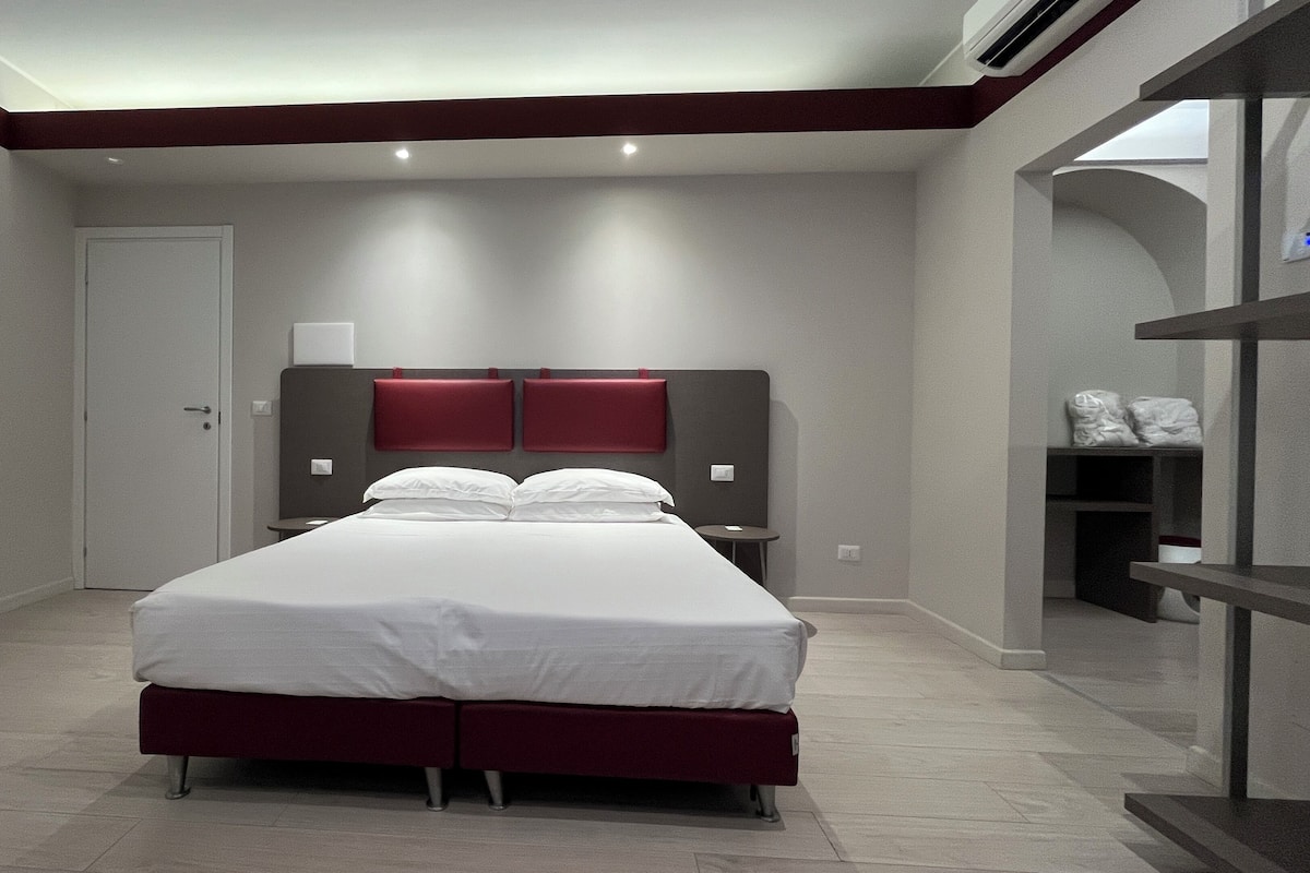 A modern hotel room with a large bed, a bedside table, a wardrobe, and a built-in shelf.