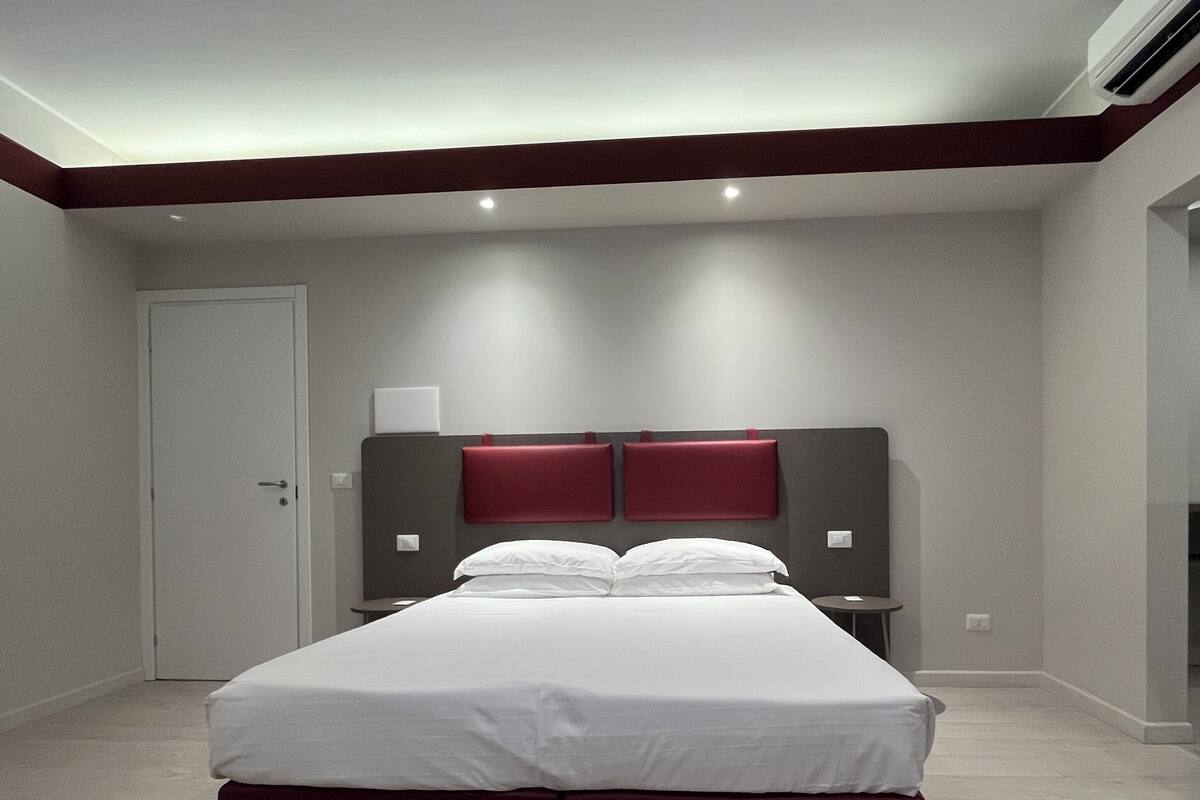 A modern hotel room with a large bed, a bedside table, a nightstand, and a wall-mounted lamp.