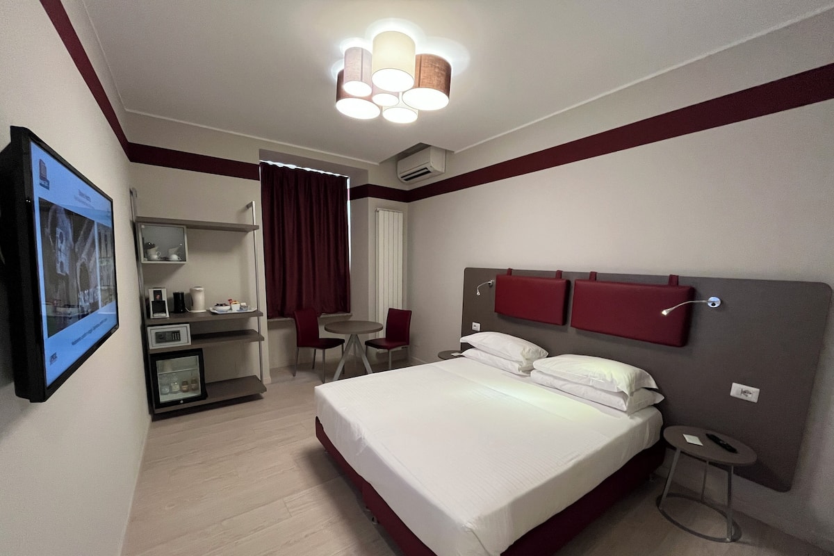 A hotel room with a bed, a desk with a computer, a chair, and a TV mounted on the wall.