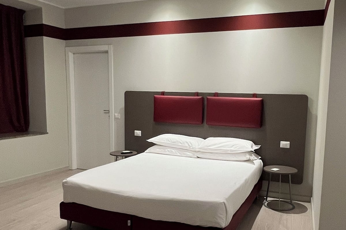 A modern hotel room with a large bed, two bedside tables, a red accent wall, and a ceiling light fixture.