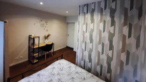 Quarto com cama, escrivaninha e cadeira. Uma parede exibe padrões geométricos e há uma porta ao fundo.