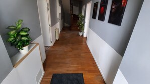 Um corredor com piso de madeira, paredes brancas e um tapete preto. Há plantas em vasos e quadros nas paredes.
