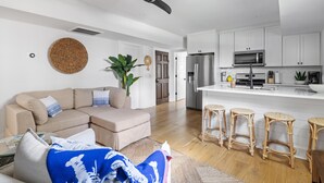 A modern living room with a beige sectional sofa, a blue blanket, and a ceiling fan. Adjacent to the living room is a kitchen with white cabinets, a stainless steel refrigerator, and a bar area with wicker stools. - A newly renovated classic Charleston home in the heart of downtown  (Charleston)