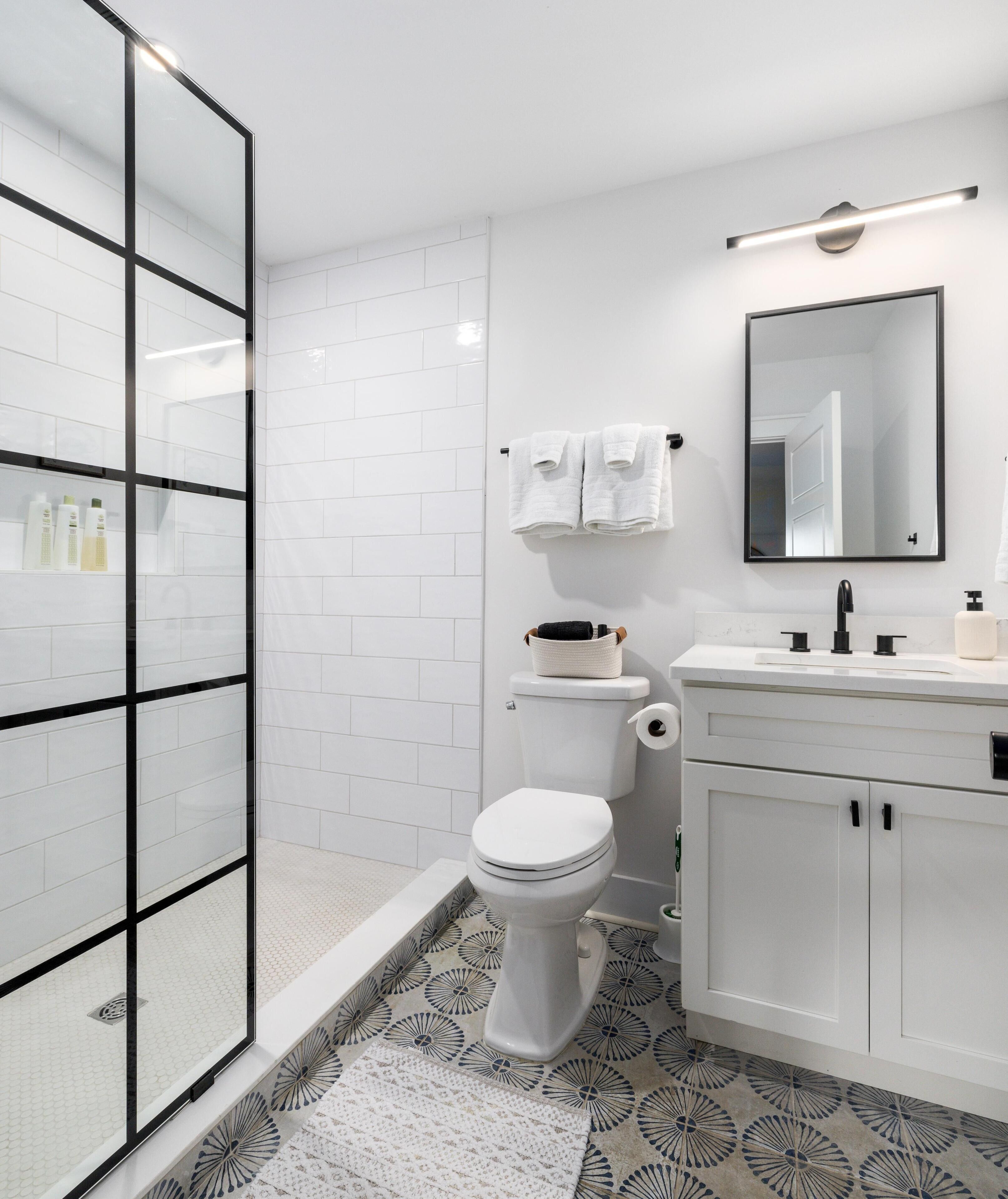 A modern bathroom with a glass shower enclosure, white tiled walls, a toilet, a sink with a mirror, and a patterned floor.