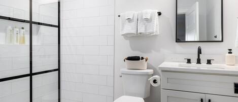 A modern bathroom with a glass shower enclosure, white tiled walls, a toilet, a sink with a mirror, and a patterned floor.