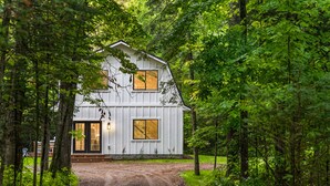 A white wooden cabin with a dirt driveway, surrounded by dense forest. - Cabin Near Banning State Park, the Munger Trail & Grindstone Lake -Dogs Welcome! (Sandstone)