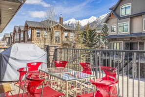 Un espacio de estar al aire libre cubierto con sillas rojas y una mesa de vidrio, con vistas a una cordillera y un edificio residencial.