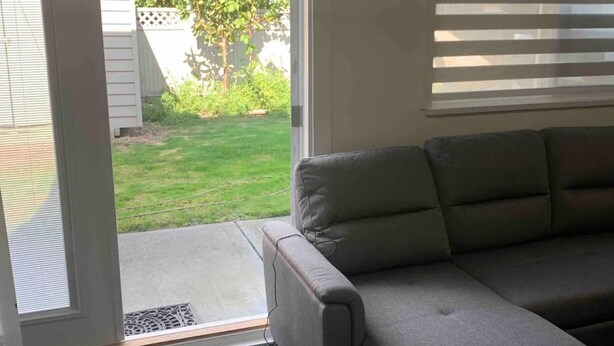 Un soggiorno con un divano angolare grigio, una porta vetrata scorrevole che dà su un giardino e una finestra con persiane.