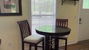 A dining area with a round table and two chairs, a window with blinds, a framed picture on the wall, and a door with a coat rack above it.
