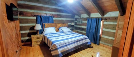 A cozy cabin bedroom with a wooden ceiling, a bed with striped bedding, a nightstand with a lamp, and blue curtains.