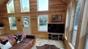 A cozy cabin interior with a leather sofa, a large cowhide rug, wooden walls, and multiple windows offering views of the outdoors. - Slow Your Roll - 2 King Suites • Near Bluff Dale Winery (Bluff Dale)